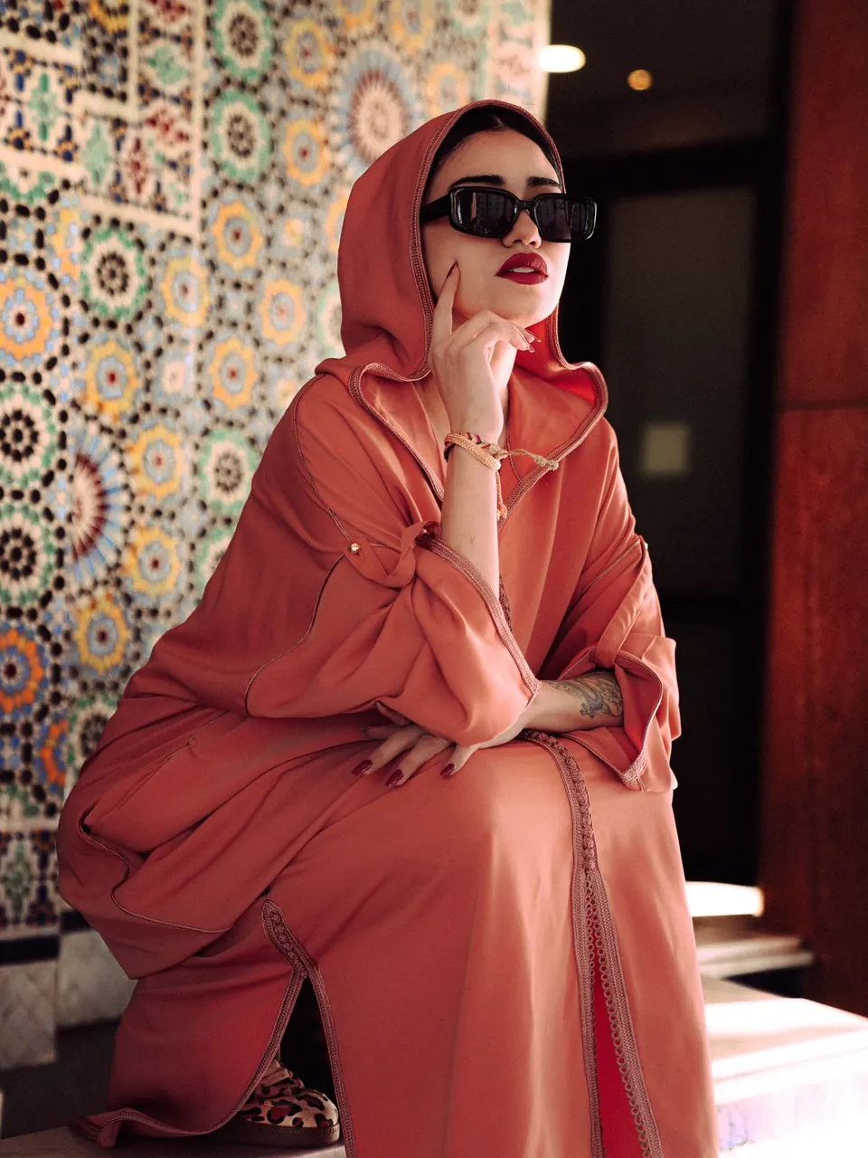 Une femme en djellaba corail et lunettes de soleil devant des zelliges