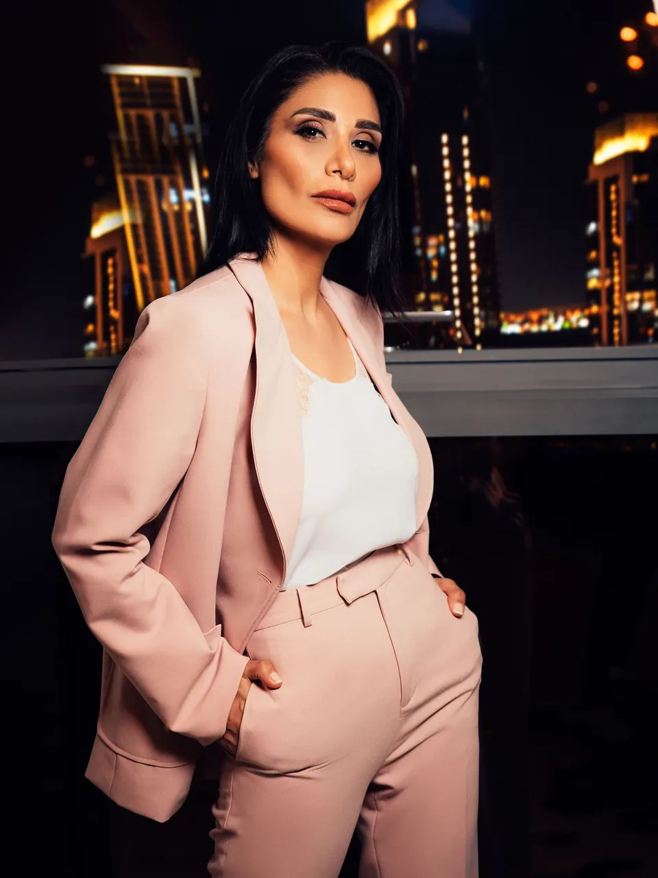 Une femme en tailleur rose blush, de nuit, devant les lumières de la ville