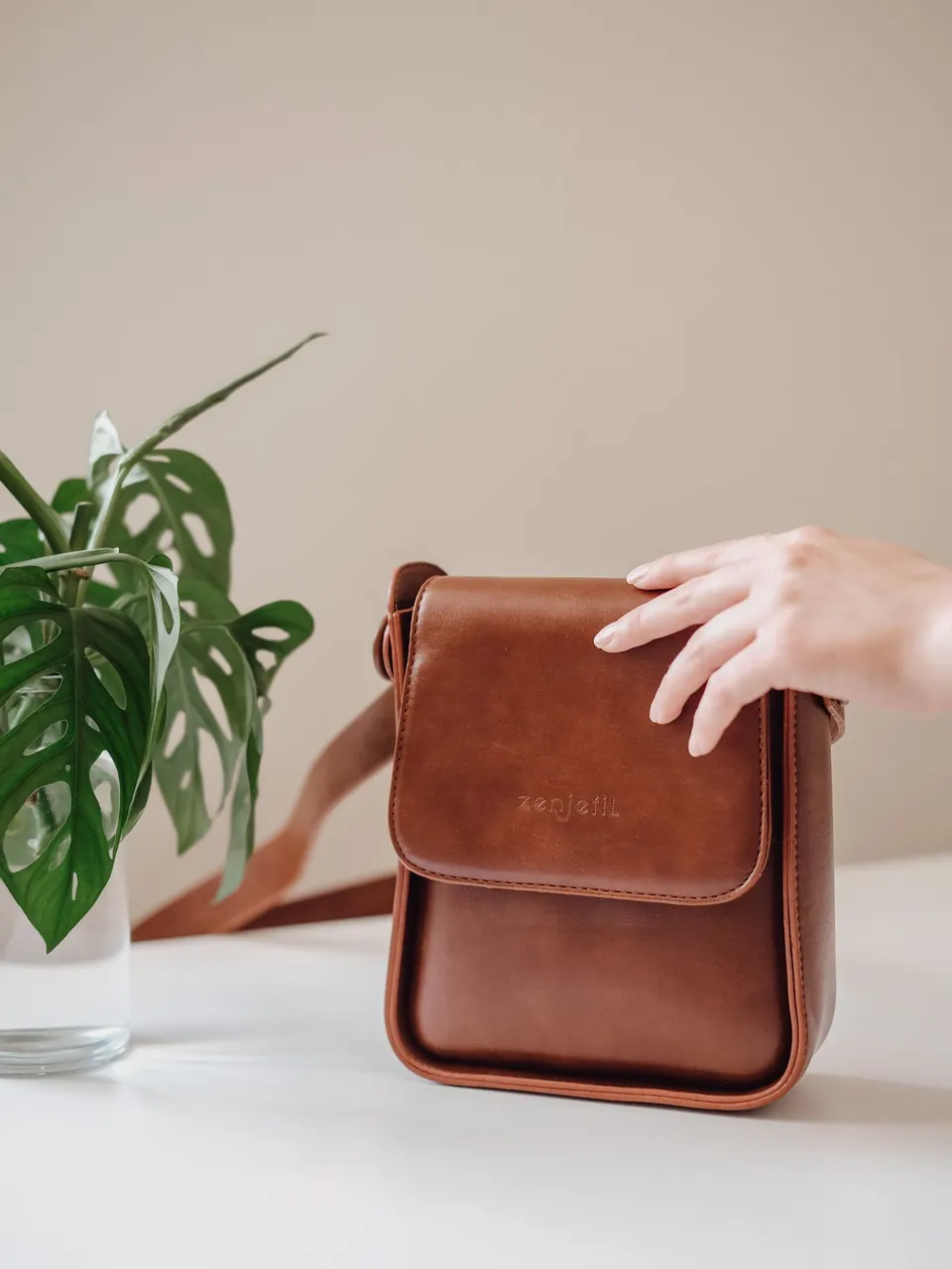 Un sac bandoulière en cuir brun posé sur une table, une main sur le rabat